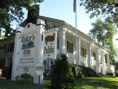 A charming white inn with a sign reading "Rider's Inn" and "Open to the Public," surrounded by greenery.