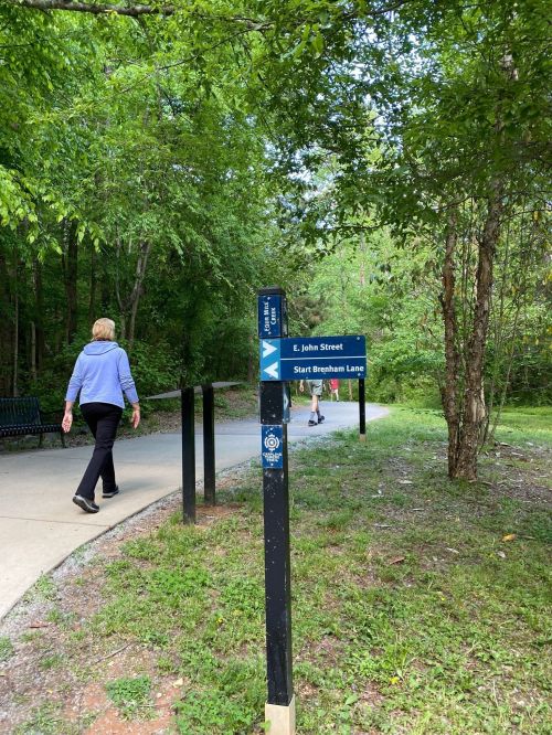 A person walks along a path with directional signs for E. John Street and Breham Lane in a wooded area.