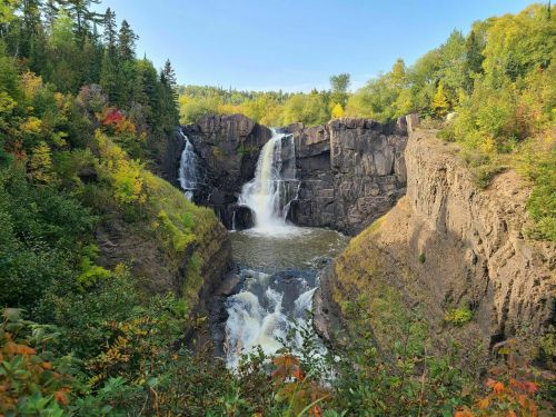 A scenic view of a waterfall cascading between rocky cliffs, surrounded by vibrant autumn foliage and clear blue skies.
