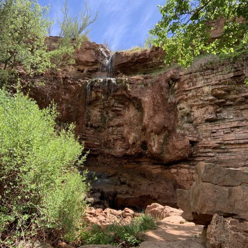 A rocky cliff with a small waterfall surrounded by greenery under a blue sky.