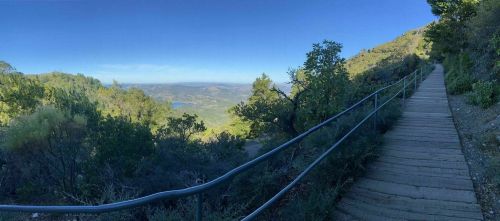 A scenic view from a wooden path, surrounded by greenery, overlooking a valley and distant mountains under a clear blue sky.