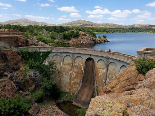 A dam with a spillway, surrounded by rocky terrain and lush greenery, overlooking a calm lake under a blue sky.