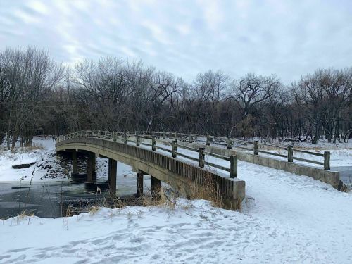 A wooden bridge arches over a snowy landscape, surrounded by bare trees and a frozen stream.