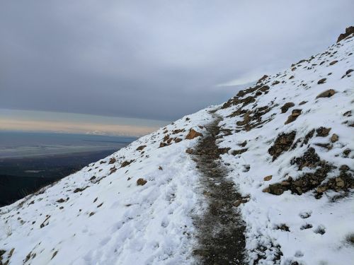 A snowy mountain path winds along a rocky slope, with a view of the ocean and cloudy sky in the distance.