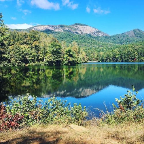 A serene lake surrounded by lush greenery and mountains, reflecting the clear blue sky and vibrant foliage.