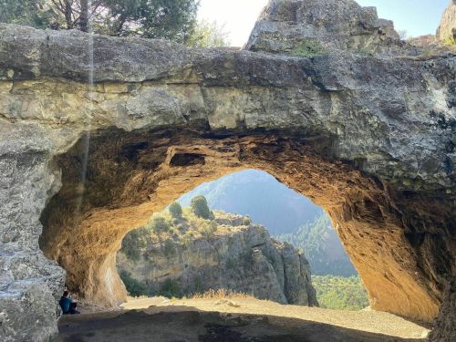 A natural stone archway frames a scenic view of mountains and trees in the background. Sunlight filters through the arch.