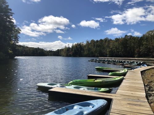 A serene lake scene with kayaks lined up on a wooden dock under a partly cloudy sky.