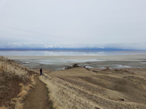 A person walks along a dirt path overlooking a vast, flat landscape with mountains in the distance under a cloudy sky.