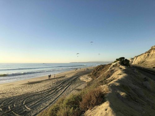 A serene beach scene with people walking along the shore and paragliders in the clear sky above.