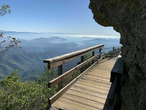 A wooden overlook path on a mountain, with a view of misty hills and a distant city skyline.