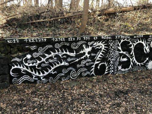 A black and white mural on a stone wall, featuring swirling patterns and abstract designs in a wooded area.