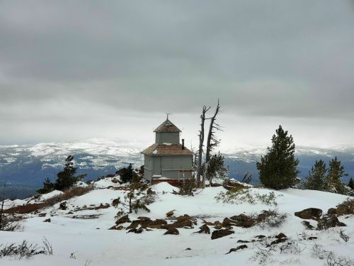 A small, weathered building on a snowy mountain peak, surrounded by rocky terrain and distant snow-covered hills under gray skies.