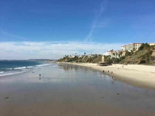 A sandy beach with gentle waves, people walking, and coastal buildings under a clear blue sky.