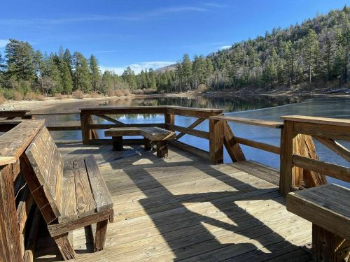 A wooden deck overlooking a calm lake surrounded by trees and mountains under a clear blue sky.
