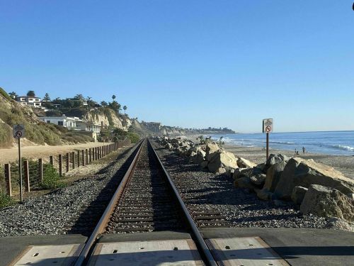 View of train tracks along a beach, with cliffs and houses in the background under a clear blue sky.