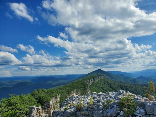 A panoramic view of mountains under a blue sky filled with fluffy clouds, with rocky foreground and lush green hills.