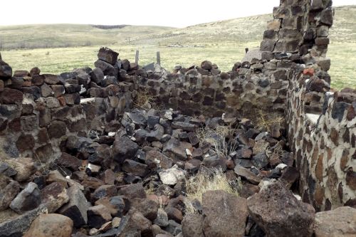 Ruins of a stone structure with scattered rocks and grass, set against a grassy landscape and distant hills.