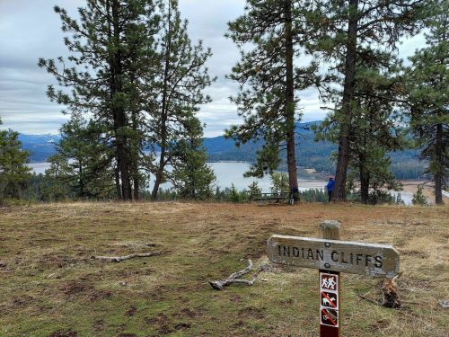 A wooden sign reading "Indian Cliffs" in a forested area, overlooking a lake and mountains in the distance.