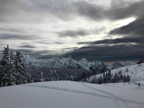 A snowy mountain landscape under a cloudy sky, with distant peaks and a few people in the foreground.