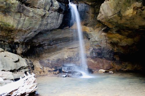 A serene waterfall cascades into a calm pool surrounded by rocky cliffs and natural stone formations.