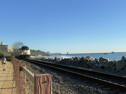 A train approaches along the coast, with a clear blue sky and a pier in the background. A person walks nearby.