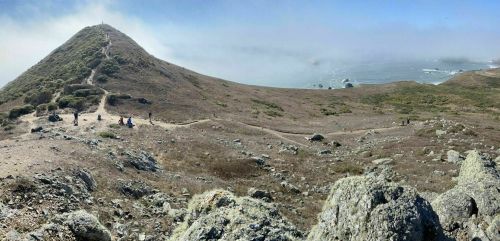 A panoramic view of a coastal landscape with a grassy hill, rocky terrain, and people walking along a path.