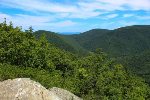 Lush green mountains under a blue sky with scattered clouds, viewed from a rocky outcrop.