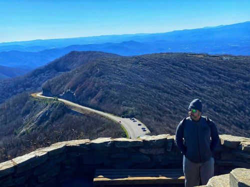 A person stands on a stone overlook, gazing at a winding road and mountains under a clear blue sky.