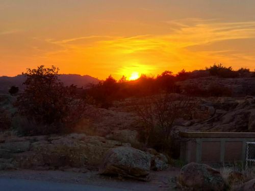 Sunset over rocky terrain with silhouetted trees and distant hills, casting warm orange and yellow hues in the sky.