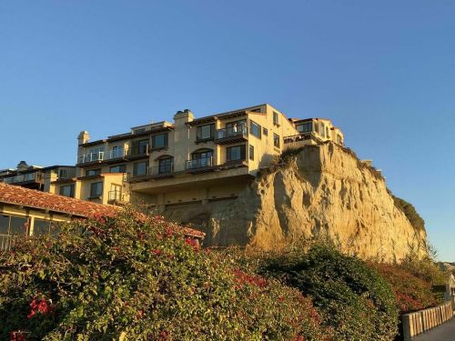 A cliffside hotel with balconies, surrounded by greenery, under a clear blue sky during sunset.