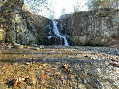 A serene waterfall cascading over rocky cliffs into a shallow stream, surrounded by trees and autumn leaves.