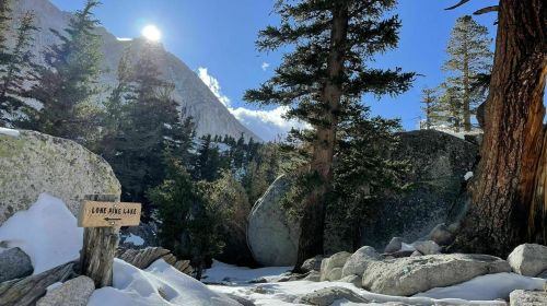 A snowy landscape with a wooden sign reading "Lone Pine Lake," surrounded by tall trees and mountains under a clear blue sky.