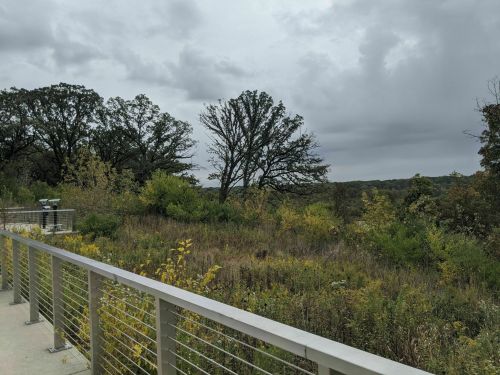 A view from a railing overlooking a lush, green landscape with trees and cloudy skies in the background.