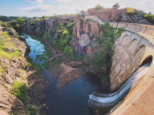 A scenic view of a dam spilling water into a rocky river surrounded by greenery under a bright sky.