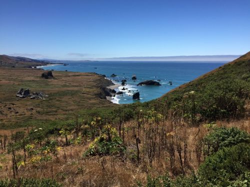 A scenic coastal view with rocky shores, blue ocean, and green hills under a clear blue sky.