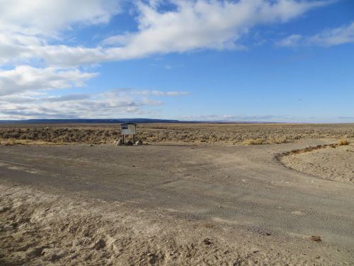 A desolate landscape with a small structure on a dirt road, surrounded by sparse vegetation and a blue sky.