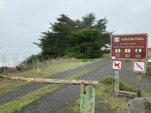 Sign for Kortum Trail, indicating trail length and elevation change, with a path leading towards the coastline and trees.