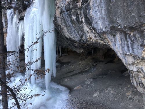 Icicles hang from a rocky overhang near a cave entrance, surrounded by snow and trees in a winter landscape.