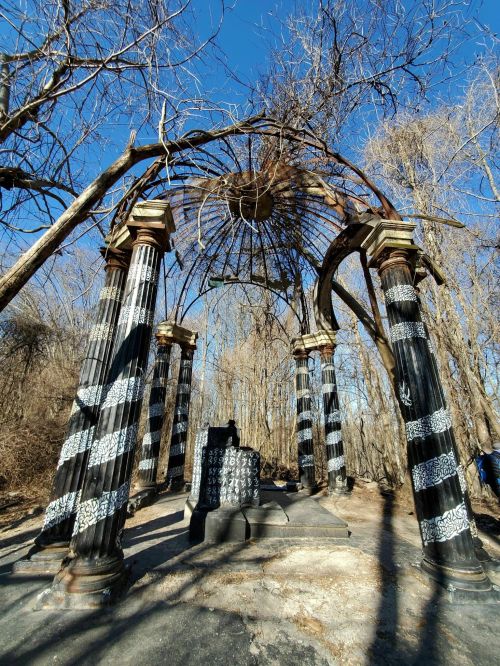 A weathered gazebo with striped columns, surrounded by bare trees against a clear blue sky.