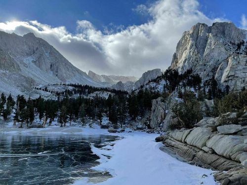 A serene winter landscape featuring a frozen lake, snow-covered rocks, and towering mountains under a cloudy sky.