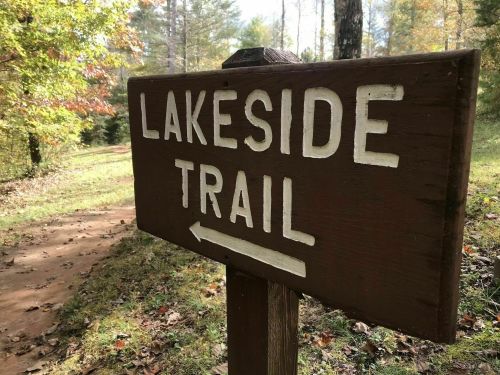 A wooden sign pointing left, reading "Lakeside Trail," with a dirt path and trees in the background.