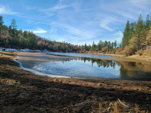 A serene lake surrounded by trees, reflecting the sky, with a sandy shore and patches of snow in the background.