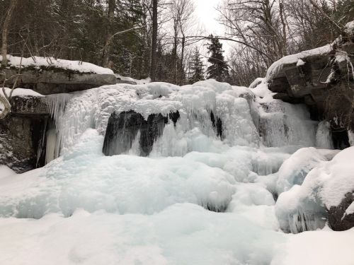 A frozen waterfall surrounded by snow-covered rocks and trees, creating a serene winter landscape.