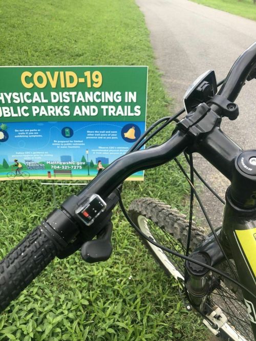 A bicycle handlebar in front of a sign about COVID-19 physical distancing in parks and trails.