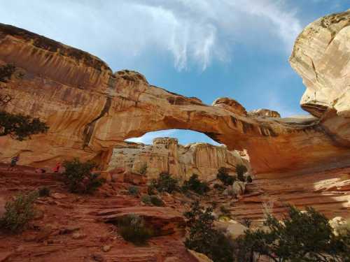 A natural rock arch rises above a desert landscape, framed by towering cliffs and scattered vegetation under a blue sky.