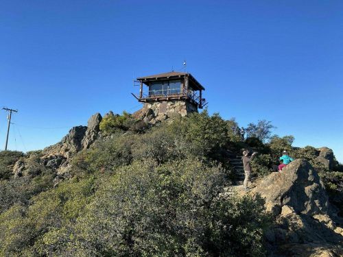 A lookout tower atop a rocky hill, surrounded by shrubs, with two people exploring the area under a clear blue sky.