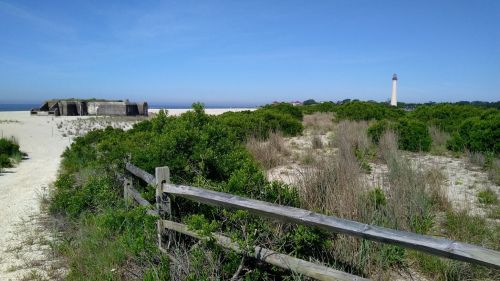 A sandy landscape with green shrubs, a wooden fence, a historic bunker, and a lighthouse in the distance under a clear blue sky.