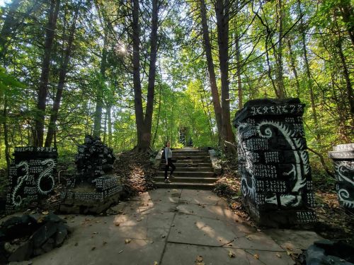 A person walks along a stone path surrounded by trees and decorated stone pillars in a lush, green forest.