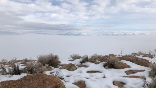 A snowy landscape with rocky terrain and low clouds, revealing distant mountains under a cloudy sky.