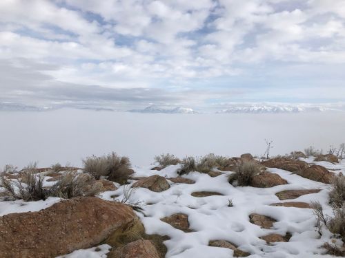 A snowy landscape with rocky terrain, under a cloudy sky, overlooking a sea of fog and distant mountains.